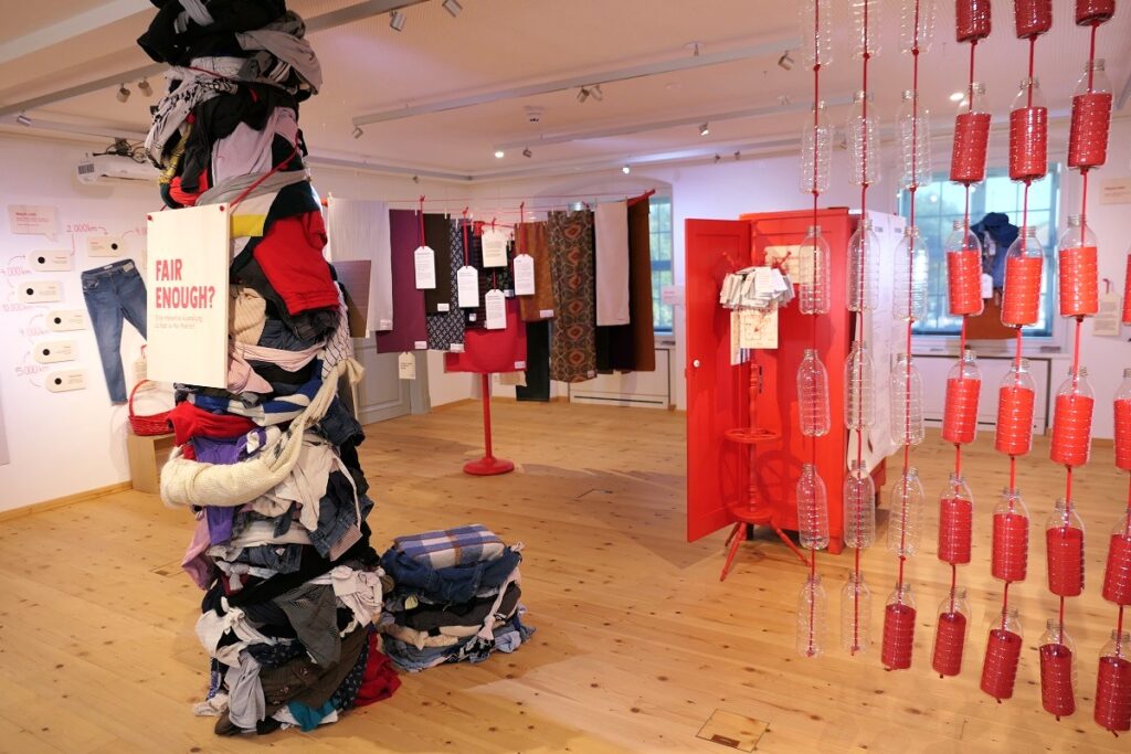Blick in die Ausstellung "fair enough". Im Vordergrund eine mit Altkleidern umwickelte Säule, daneben eine Vorhang aus Pet-Flaschen, durch den man in den Raum blickt.
